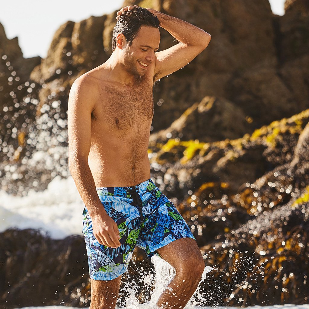 Man wearing blue tropical botanical boardshorts standing in ocean water near rocks, premium beach and surf swimwear